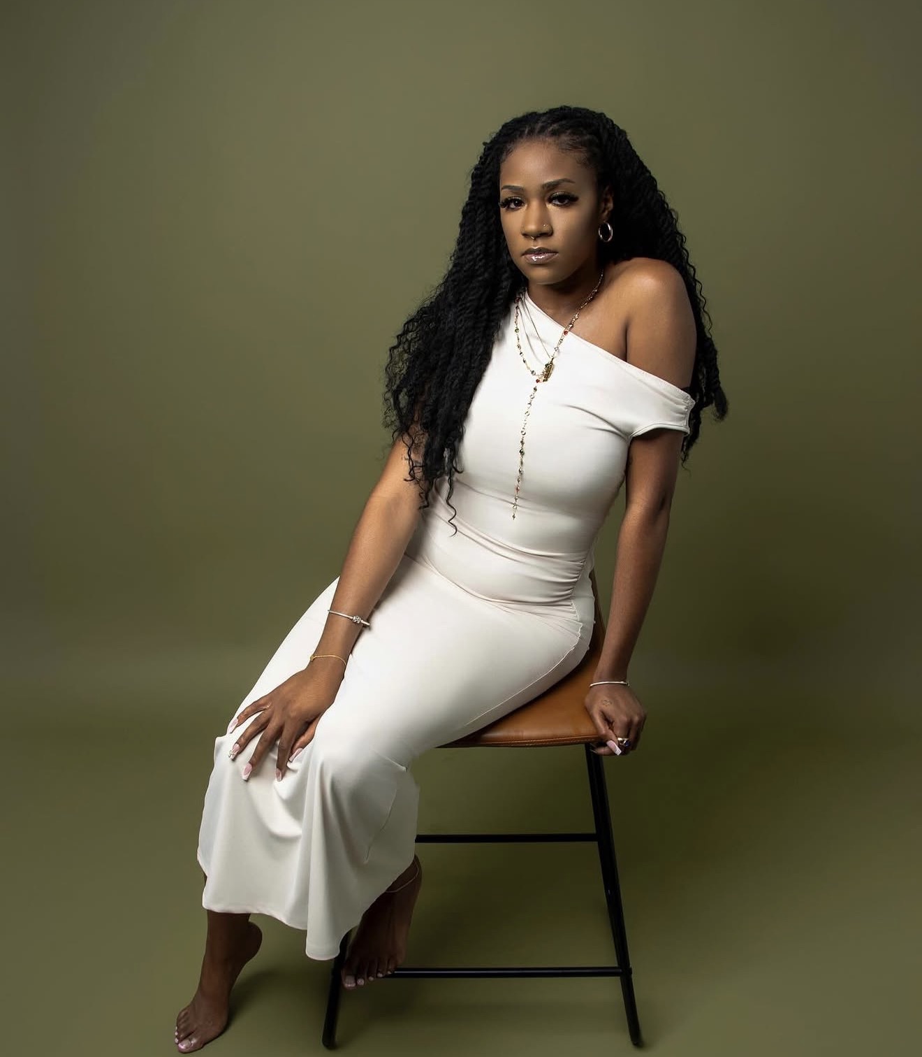 Drizzy, the stylist behind Loc Therapy, seated on a stool in a cream dress against an olive studio backdrop, her long locs falling over one shoulder