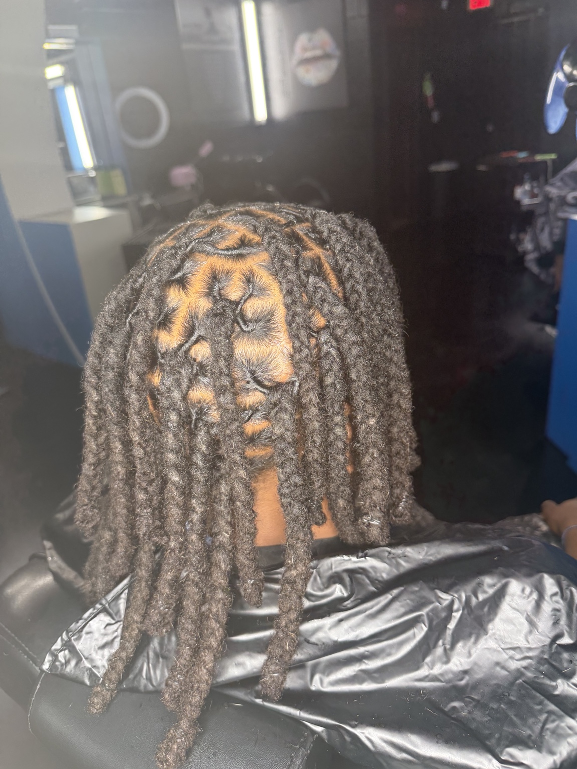 Client caped in the chair in front of the salon mirrors, a freshly retwisted crown parted in a clean grid