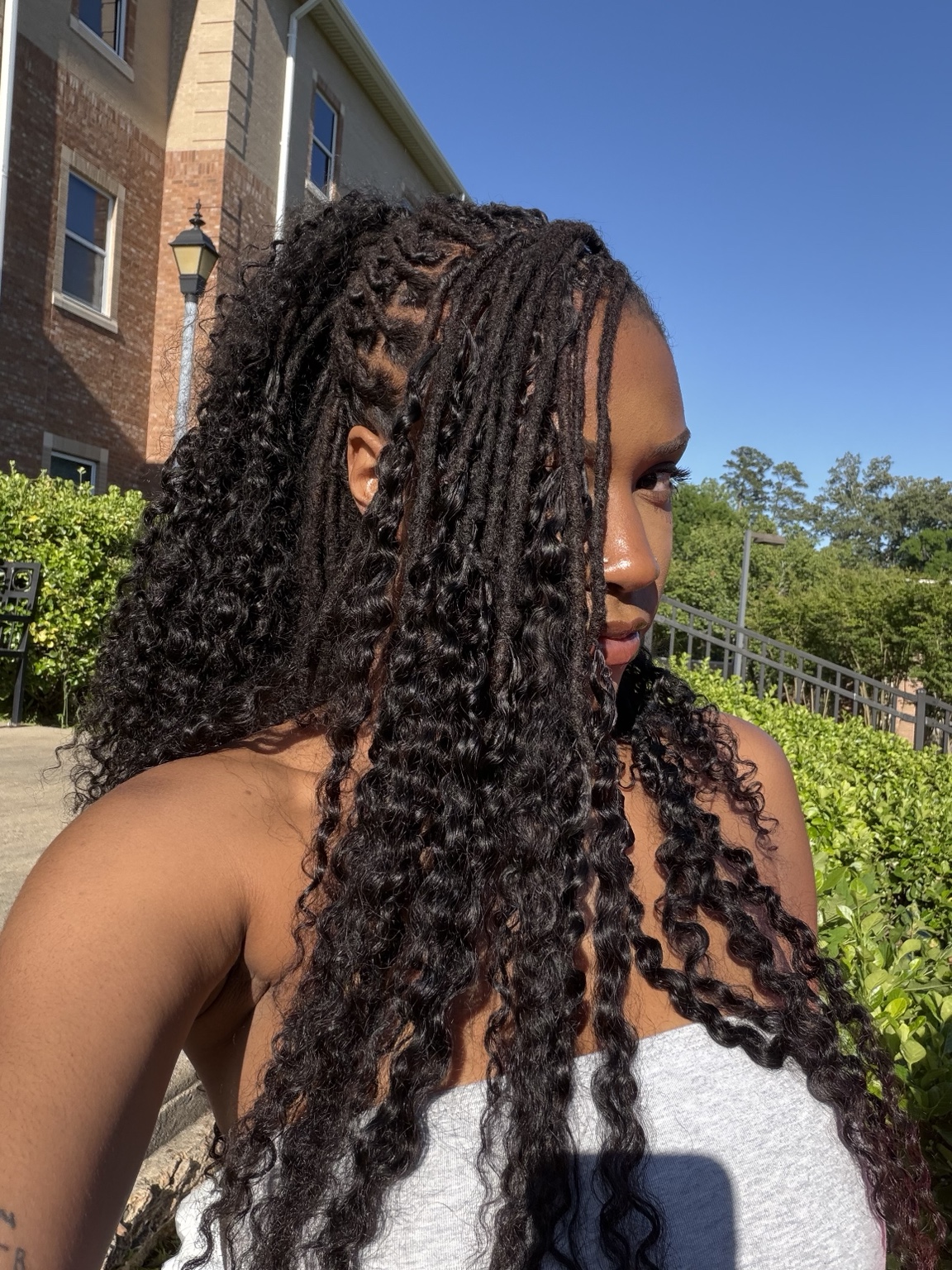 Client outdoors in bright afternoon light, turned away from the camera and looking over her shoulder, wearing long curly loc extensions that fall past her shoulders against green hedges and a brick wall