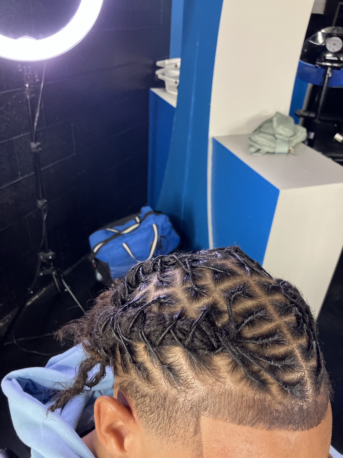 Client in the salon chair under the ring light, his short locs retwisted into a diamond parting pattern above a clean taper